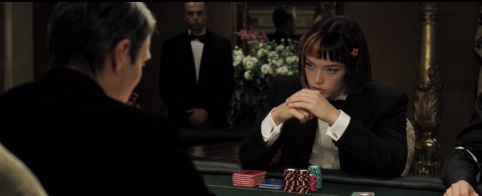A scene from a poker movie showing a young woman with a black bob haircut and an 'A' playing card in her hair, sitting across the green baize from an older man in a tuxedo setting.