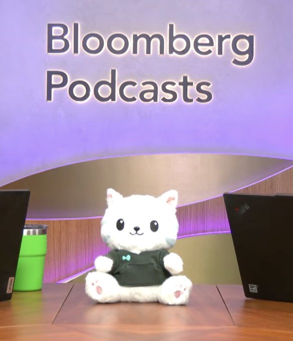 A cute white bear mascot plush sits at a desk in front of a "Bloomberg Podcasts" backdrop, representing the podcast episode where Tracy Alloway and Eric Balchunas discuss how Jeff Bezos's Jeff is set to house finance, with the post mentioning Hyperliquid.