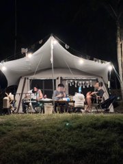 A group sits around a candlelit table under a starlit white tent at night, with two people focused on their laptops—one showing charts and tickers like "BTC"—proving that even in nature, the crypto grind doesn't sleep.