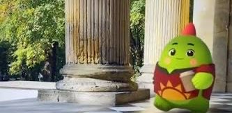 A green cartoon cactus character wearing a red and yellow patterned poncho and a small red hat, standing near stone columns in a sunny courtyard.