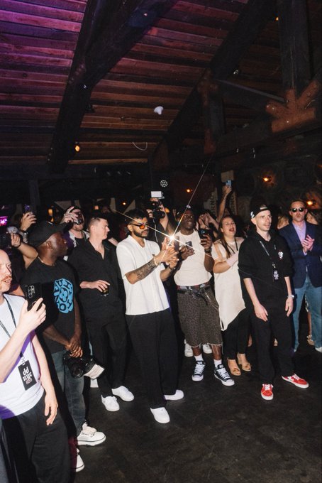 A nighttime crowd photo shows people gathered in a venue with wooden beams, many holding up phones to record, with one person in a white shirt and sunglasses prominently capturing the moment.