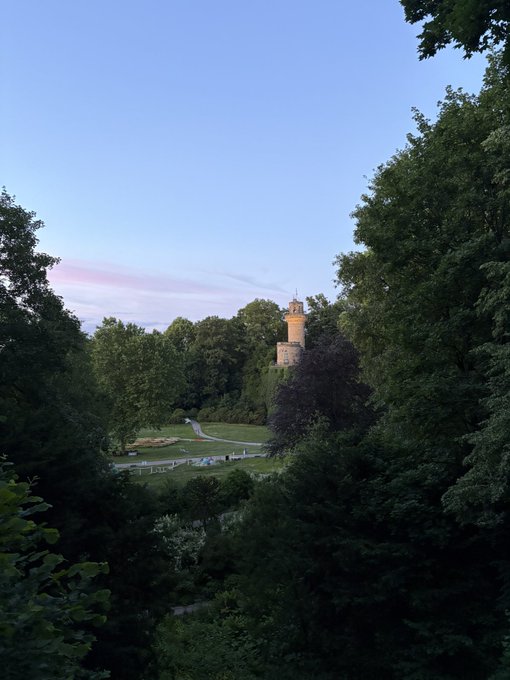 The image shows a beautiful park with trees, grass, and a large tower that resembles an old castle or a church steeple. The sky above has a pinkish hue, creating a picturesque scene. In the foreground of the photo, there is a path leading towards the tower, inviting visitors to explore its history and architecture.