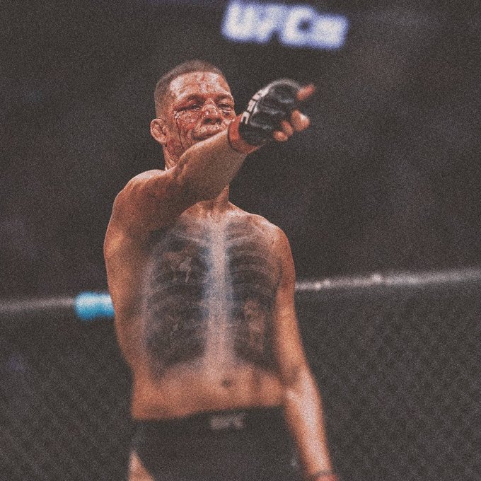 A bloodied UFC fighter with chest tattoos points toward the camera inside the octagon during a match.