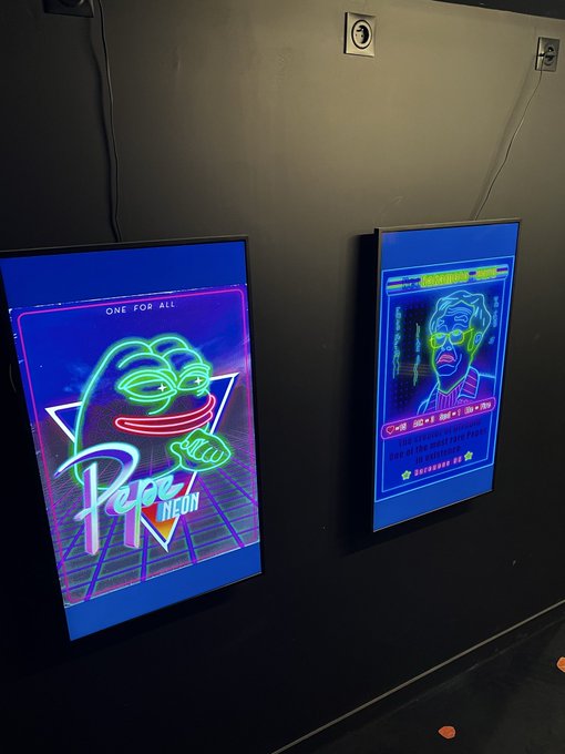 Two glowing neon-style digital screens mounted on a dark wall, displaying retro-futuristic graphics including a palm tree, a character face, and text reading 'ONE FOR ALL' and 'Pepe Neon'.