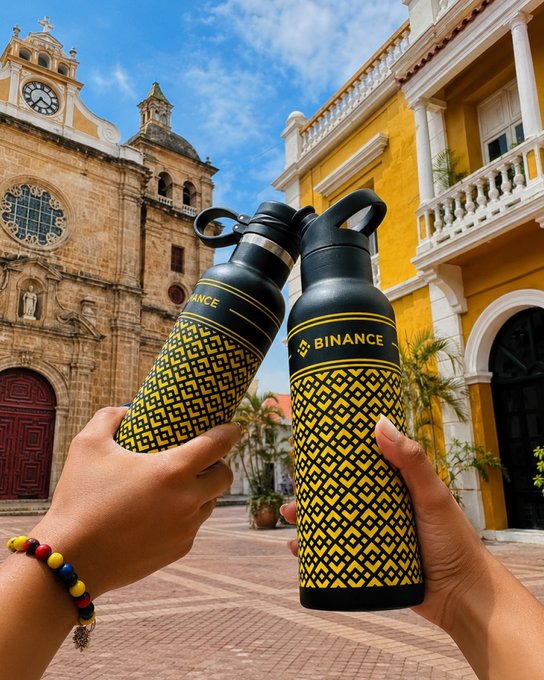 Two hands clink branded water bottles—one labeled "BINANCE" and the other partially showing "ANCE"—in a toast against the backdrop of a historic yellow colonial building and stone church, capturing a travel moment between crypto enthusiasts with the caption "just a solid duo."