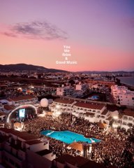 An aerial photograph of a crowded resort or event venue at sunset, featuring a large swimming pool and beach setup with the text 'This is Sant Llorenç' overlaid.