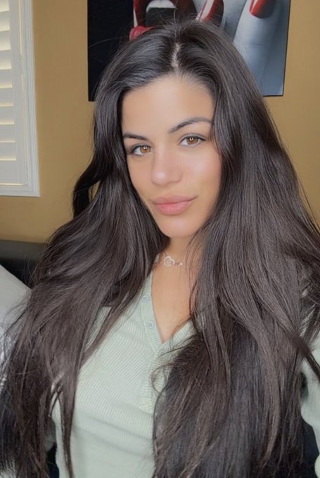 A selfie-style portrait of a young woman with long, wavy dark brown hair wearing a light green ribbed top and a small pendant necklace, with a framed artwork visible on the wall behind her.
