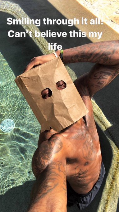A shirtless, tattooed man in a pool wearing a brown paper bag with eye holes over his head, with text overlay reading 'Smiling through it all! Can't believe this my life'.