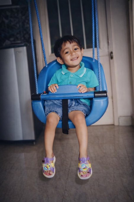 A joyful photo of a young smiling child sitting in a blue plastic swing seat, wearing a teal polo shirt, denim shorts, and colorful cartoon sandals.