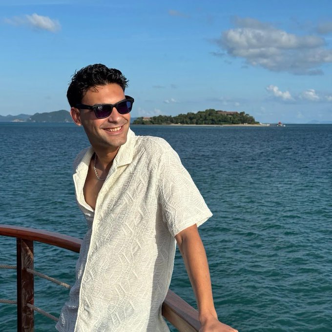 A photograph of a smiling man wearing sunglasses and a white textured short-sleeved shirt, posing by a railing with the ocean and a tropical island in the background.