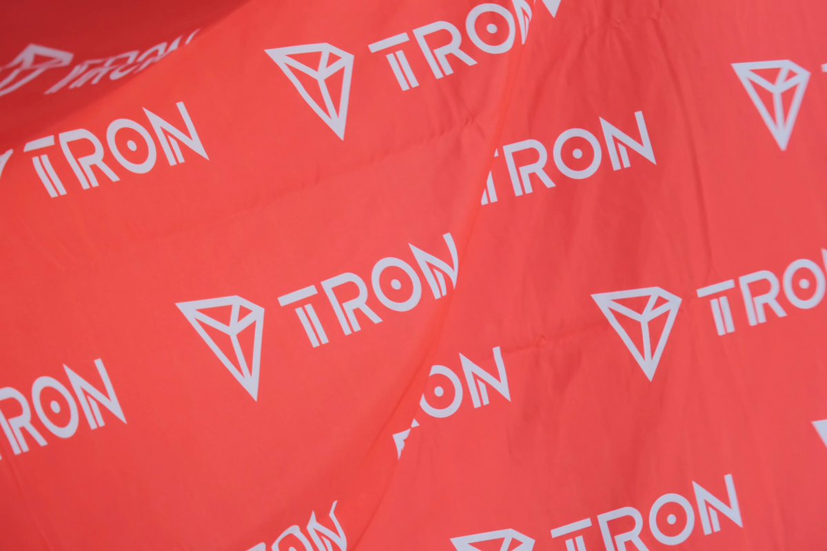 The image features a close-up view of a red fabric that has white logos on it. The text "TRON" can be seen, indicating that this might be an advertisement or promotional material related to the TRON Academy. The focus is on the vibrant red color and the white logos, which are prominently displayed in the foreground.