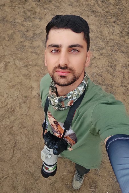 A man with a beard wearing a green t-shirt and patterned scarf takes a selfie while holding a DSLR camera with a large telephoto lens, standing on a dirt path.