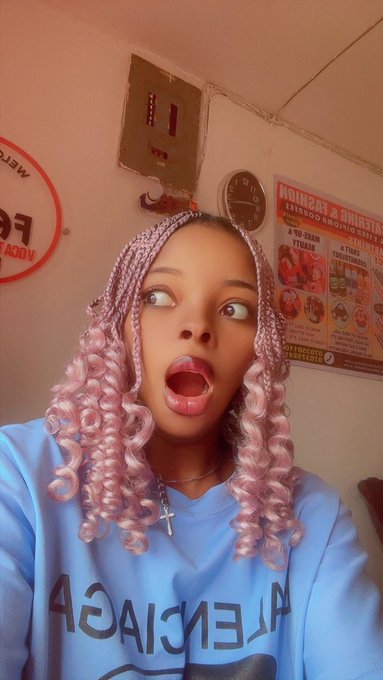 A young woman with pink and purple braided hair takes a selfie with an open-mouthed expression, wearing a light blue Balenciaga sweatshirt in what appears to be a kitchen.
