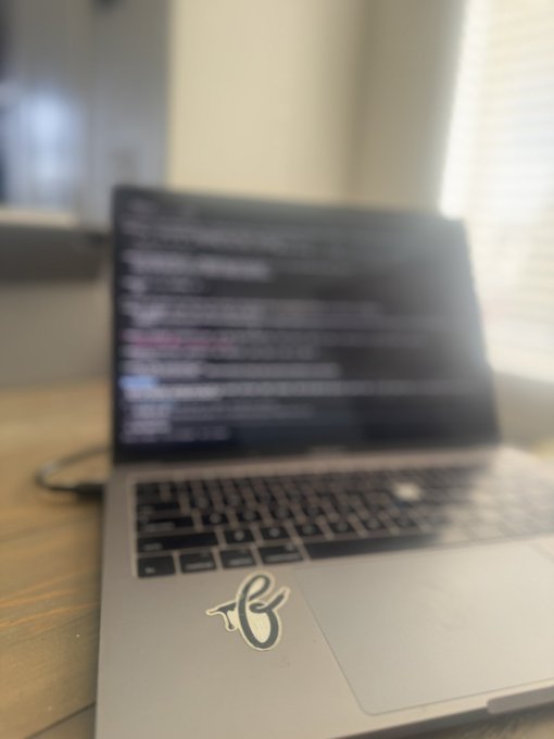 A MacBook laptop on a wooden desk displays lines of code on its screen, with a sticker featuring a stylized "B" logo on the palm rest, symbolizing the process of "building" software in progress.