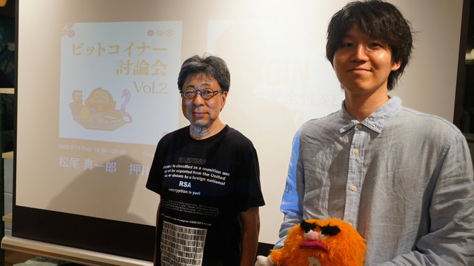 Two men stand in front of a presentation slide titled "ビットコイナー討論会 Vol.2" (Bitcoiner Discussion Vol. 2) with the date "2024.8.13 THU 18:30-22:00" and the name "松尾真一郎" visible, while the younger man on the right holds a stuffed lion toy. The setup suggests a Bitcoin meetup or discussion event, contextualizing the post's commentary about Bitcoin's role as payment versus settlement.