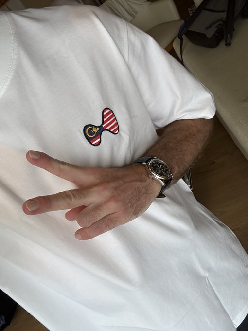 A person wearing a white shirt with a Malaysian flag patch makes a peace sign while sporting a luxury watch, proudly repping their Malaysian roots in the crypto community.