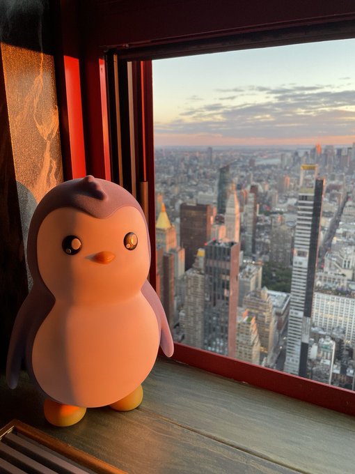 A cute penguin figurine sits on a windowsill overlooking a sprawling city skyline at sunset, bathed in warm orange light. The scene ties to the $PENGU token, fittingly featuring a penguin mascot enjoying a high-rise view over the cityscape.