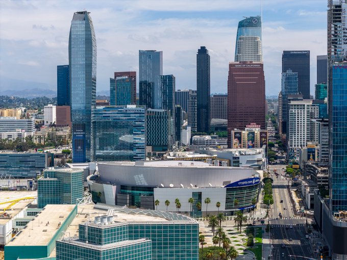 An aerial view of the Los Angeles downtown skyline featuring Crypto.com Arena, Bank of California, and other skyscrapers under a partly cloudy sky.