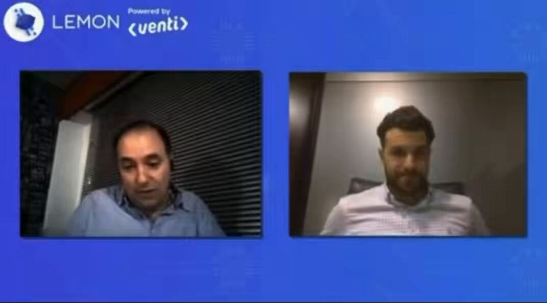 The image shows two men sitting next to each other with their faces displayed on a computer screen, likely during an online meeting or video call. The background of the image features a blue wall with white text that reads "Lemon powered by Venti". One man is wearing a blue shirt and the other has a beard. They appear to be engaged in conversation about Bitcoin, as indicated by their expressions and body language.