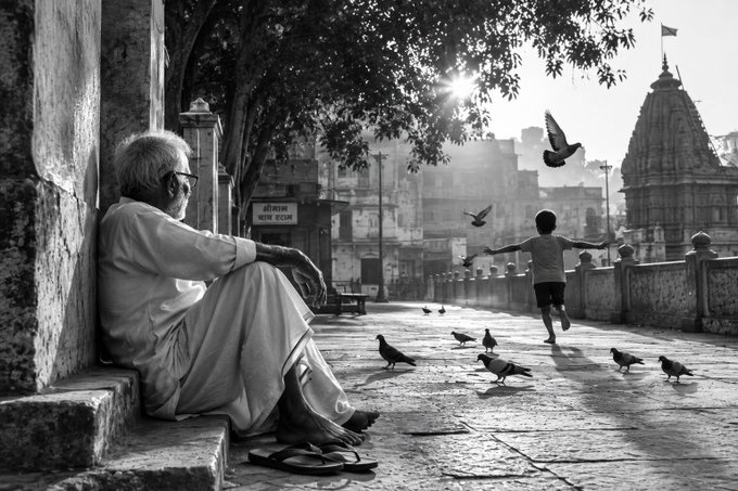A black and white street photography scene of an elderly man sitting on stone steps watching a child run with outstretched arms among pigeons, with a temple tower visible in the background.