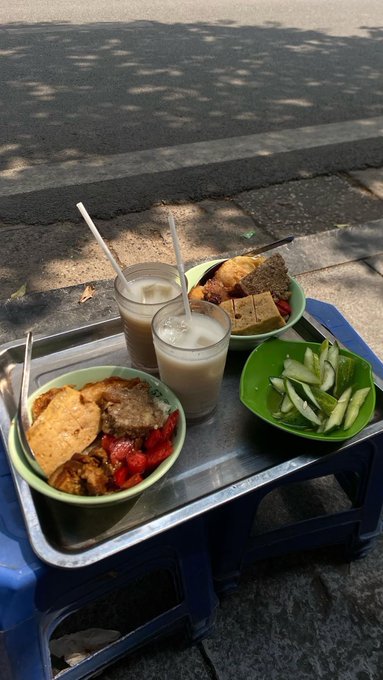 A street food meal is served on a small blue plastic table, featuring bowls of tofu, vegetables, and meats alongside cucumber slices and two glasses of a milky beverage with straws.