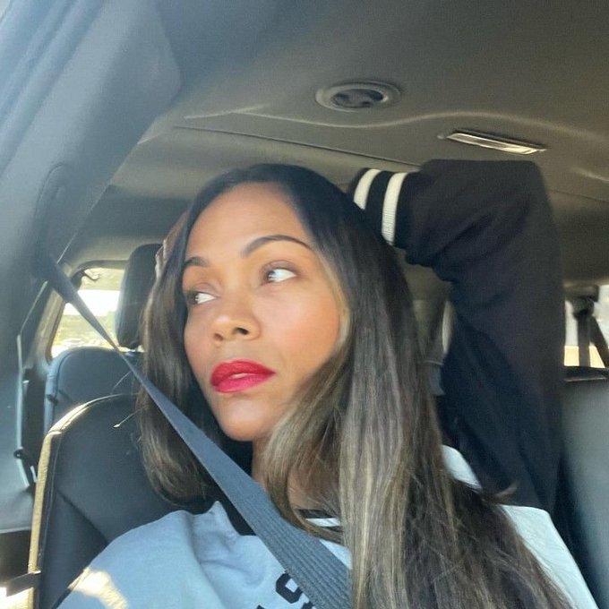 A woman with long highlighted brown hair and red lipstick poses for a selfie inside a car, gazing off to the side with one arm raised behind her head, wearing a black and white varsity-style jacket over a light blue shirt while secured by a seatbelt, capturing a relaxed morning moment that complements her grateful Sunday greeting to her Web3 community.