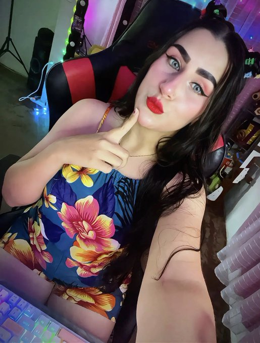 A woman with dramatic makeup takes a selfie while seated in a gaming chair, wearing a floral top.