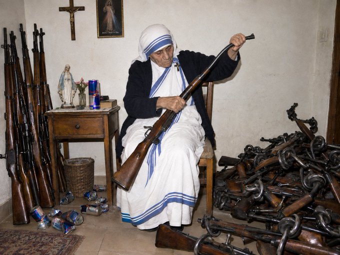 A woman dressed as Mother Teresa sits on a wooden chair, examining a rifle, surrounded by many other rifles and piles of chains in a room with religious statues and a crucifix on the wall.