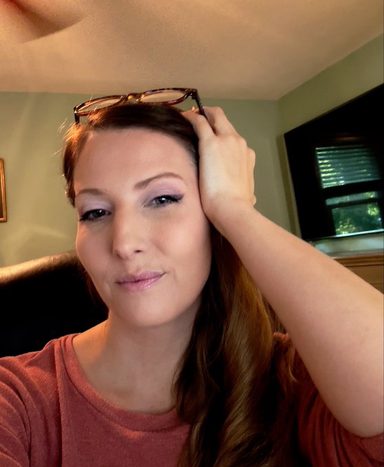 A woman with long auburn hair and sunglasses perched on her head poses for a selfie before filming, wearing a rust-colored top and natural makeup, looking camera-ready for her podcast appearance with @UseArtisan.