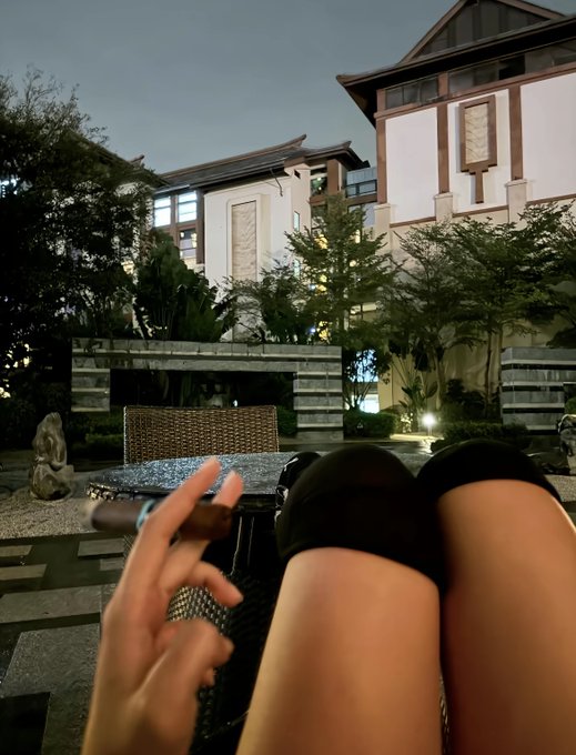 A first-person view showing legs in black shorts and a hand holding a cigar, with an Asian-style building featuring traditional architecture visible in the background.