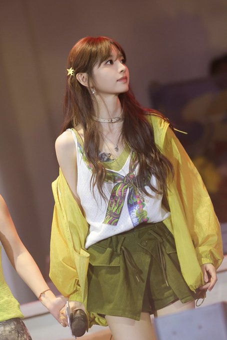 A young woman with long brown hair, wearing a star hair clip, white top, yellow sheer jacket, and olive green shorts, poses at what appears to be an event.