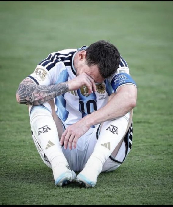 Lionel Messi in an Argentina #10 jersey sits on the grass in emotional pain, rubbing his face after a match.