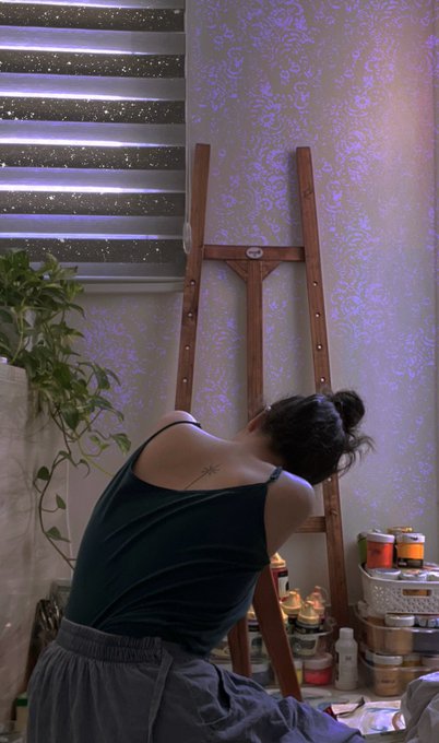 A person sits before a wooden easel painting at a table filled with supplies, illuminated by purple projected patterns on a damask wallpaper behind them.