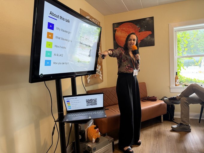 A woman giving a presentation in a casual room, pointing at a large screen displaying 'About this talk' with slide topics like 'Why Wavelength', while holding a microphone and standing next to a laptop showing a QR code.