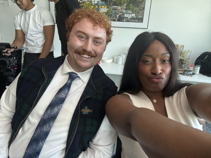 A selfie of a smiling man with red hair and a mustache wearing a plaid vest over a button-up shirt, sitting next to a woman in a white top, with another person in the background.