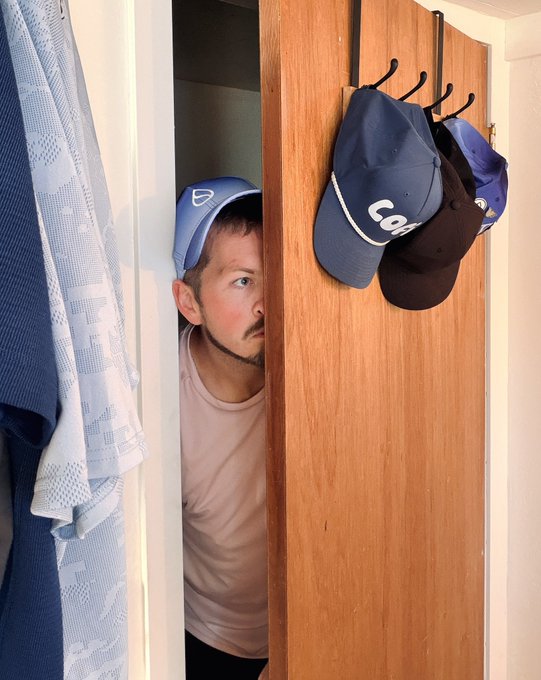 A man wearing a blue cap peeks out from behind a partially open wooden door, with several baseball caps hanging on hooks on the door.