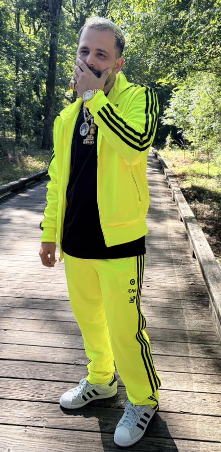 A man poses on a wooden boardwalk in a neon yellow Adidas tracksuit, with a "Bored Ape Yacht Club" graphic tee visible underneath the open jacket, playfully covering part of his face while showcasing his jewelry. The post references Bored Ape NFTs, and his tee featuring the BAYC logo directly connects his bold streetwear look to the "Feeling Apeish on this Sunday" theme.