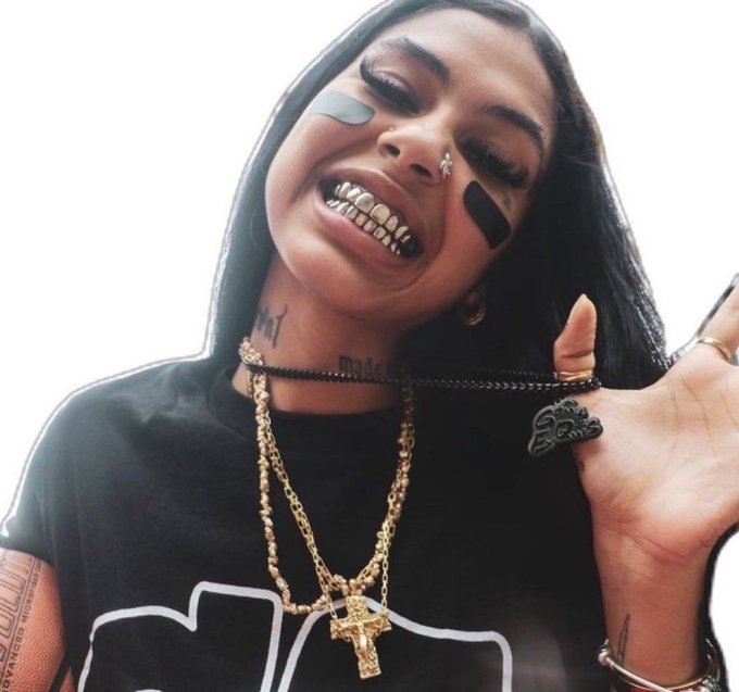 A smiling woman with face tattoos, grills, a nose ring, and multiple gold chains including a cross pendant, giving a hand gesture.