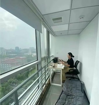 A person working on a laptop at a desk on a high-rise balcony overlooking a cityscape.