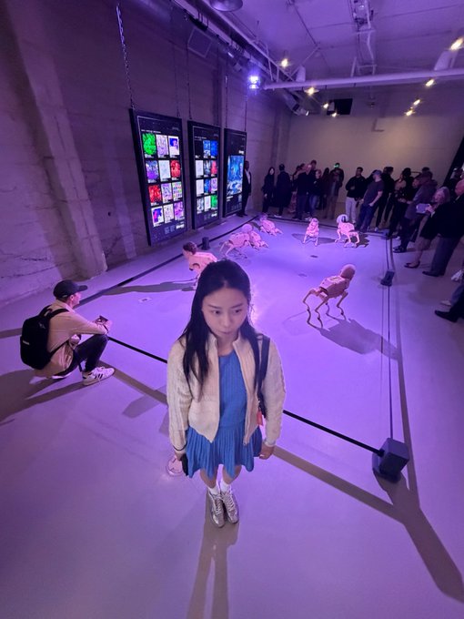 An art installation bathed in purple light features a young woman standing in the foreground, while robotic skeletal figures perform on the floor and visitors observe digital artwork displayed on vertical screens in the background.