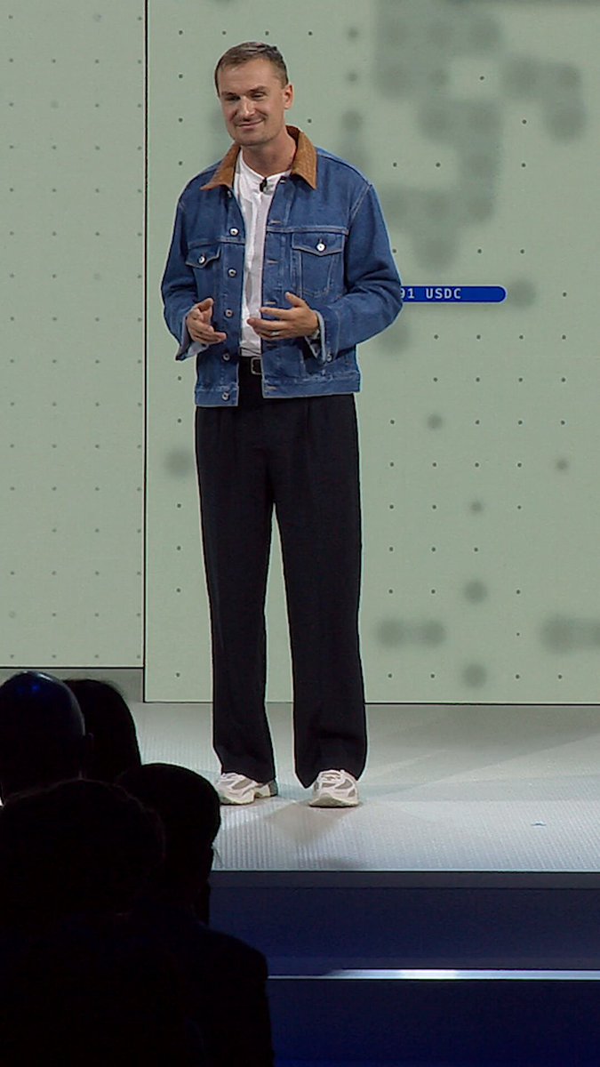 The image features a man standing on stage, wearing a blue denim jacket and black pants. He appears to be giving a speech or presentation, as indicated by his body language and the fact that he is addressing an audience. The background of the image shows a green wall with white dots, which could potentially represent a screen or projector used for displaying information during the event.