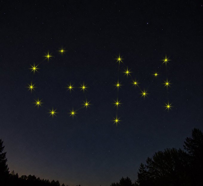 A nighttime photograph of a starry sky where illuminated drones or lights form the word 'GM' above a silhouette of trees.