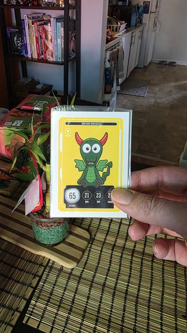 A hand holds up a holographic trading card labeled "GREEN DRAGON" with stats showing 65 stamina, 21 dribbling, and 23 defense, set against a home background with a plant and bookshelf.