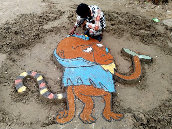 An artist kneels on the ground, carefully detailing a vibrant sand sculpture of an orange orangutan-like character wearing a blue shirt, with colorful striped tentacles extending from its sides. The whimsical creation appears to be fan art created to support "OranguNation," reflecting the creator's enthusiastic dedication shown in the post.