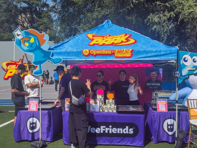 An outdoor event booth branded with 'OpenSea JUNKIES' and 'ThreeFriends' logos, featuring blue mascot characters, a blue canopy, and several people gathered around merchandise displays on purple-draped tables.