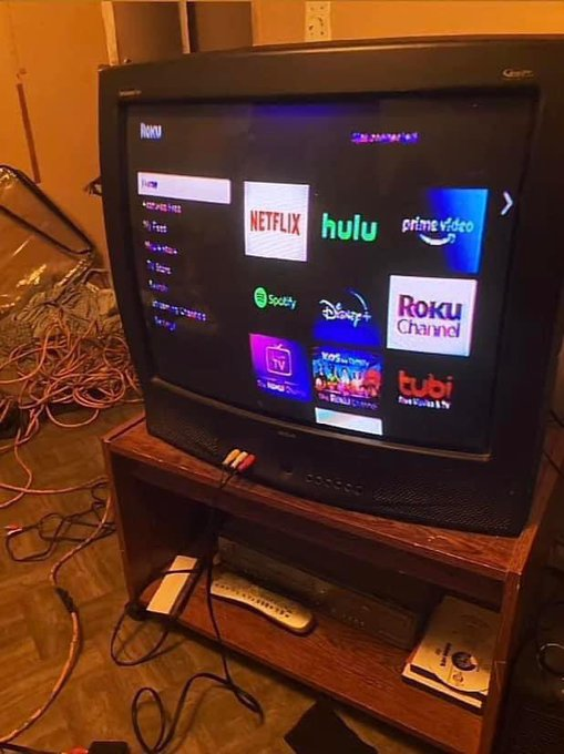 A CRT-style TV displays the Roku home screen with apps like Netflix, Hulu, Prime Video, Spotify, Disney+, and Tubi, resting on a wooden stand with tangled cords around it—symbolizing how crypto gains may have funded a new entertainment setup.