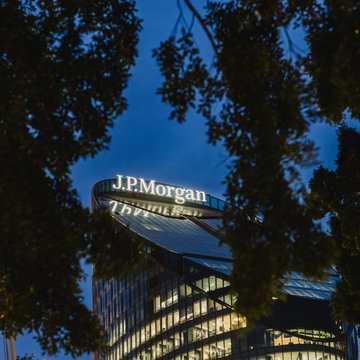 The illuminated "J.P.Morgan" sign glows atop a modern glass skyscraper against a twilight blue sky, framed by dark tree silhouettes. The image visually represents JPMorgan, the financial institution at the center of the news about its decision to drop Polymarket as a banking client while still eyeing a potential IPO underwriting role.