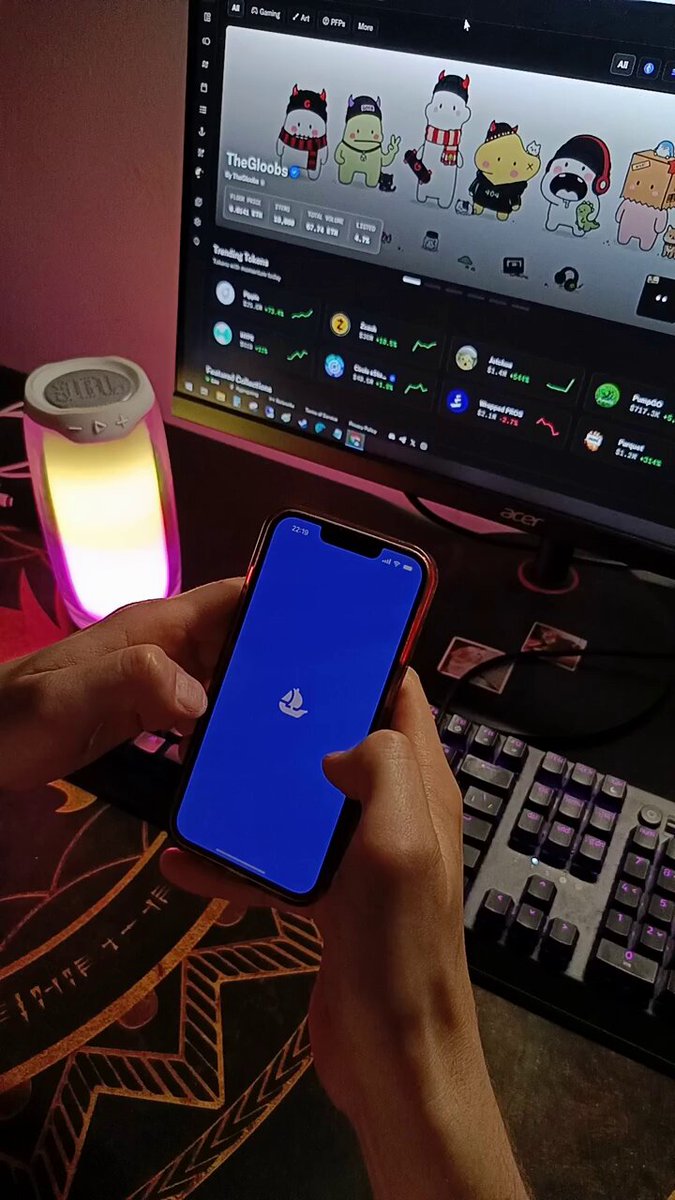 The image shows two hands holding a blue smartphone displaying an app icon, possibly a social media or gaming app like Facebook or Minecraft. The person appears to be using their phone while sitting at a desk with a computer monitor nearby. The background of the image also includes a keyboard and mouse on the desk, suggesting that this might be a workspace for someone who spends time on their phone.