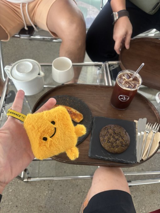 A first-person view showing a hand holding a small yellow plush toy, with a Binance-themed wristband, over a tray containing a cookie, coffee, and tea at a cafe.