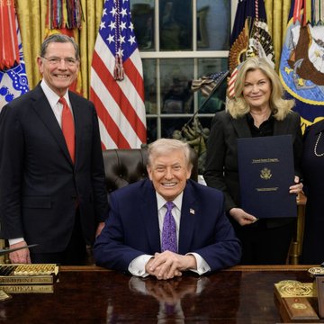 President Donald Trump is seated at the Resolute Desk in the Oval Office, smiling broadly with his hands clasped, flanked by two officials—a man in a dark suit with a red tie on the left and a woman holding a dark-bound document on the right—with American flags displayed prominently in the background. The image is being used to lend an air of official presidential gravitas to a social media post calling on followers to "like" if they want Congress to pass a Strategic Bitcoin Reserve bill, exploiting the executive setting to bolster the cryptocurrency advocacy message.